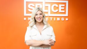 Ellie Bakker standing in front of an orange wall with the Splice Marketing logo on it. She is smiling and has her arms crossed.
