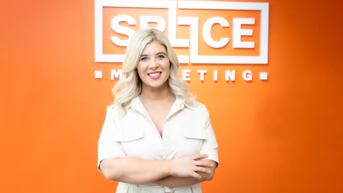 Ellie Bakker standing in front of an orange wall with the Splice Marketing logo on it. She is smiling and has her arms crossed. Healthcare marketing professionals.