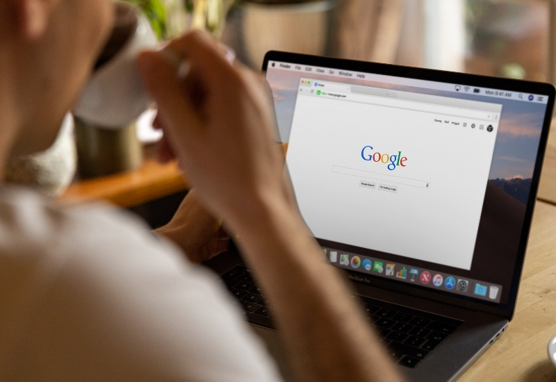 A person sits at a desk drinking from a cup while using a laptop with the Google search homepage open on the screen.