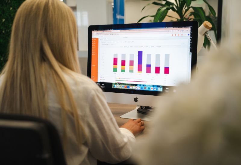 Person seated at a desk in a modern open-plan office, working on two computer monitors with plants and office furniture in the background.