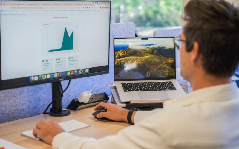 Person working at a desk using a desktop monitor displaying analytics data and a laptop, reviewing a dashboard with a rising graph on the screen.